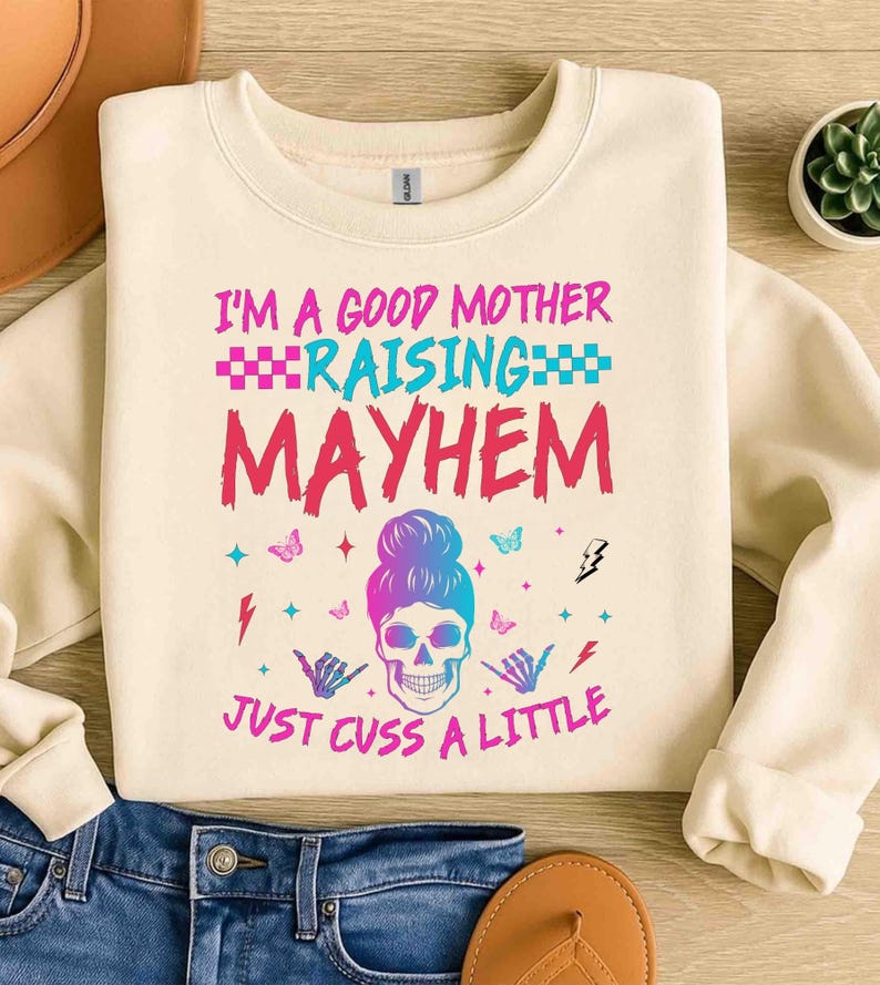 May include: Cream-colored sweatshirt with the text "I'M A GOOD MOTHER RAISING MAYHEM JUST CUSS A LITTLE" in pink and blue. The design includes a skull with a bun, butterflies, and hand gestures.