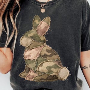 Può includere: T-shirt grigio scuro con un design a coniglio patchwork mimetico. Il coniglio è realizzato con toppe di tessuto verde, marrone e beige. La maglietta ha girocollo e maniche corte, perfetta per un look casual.