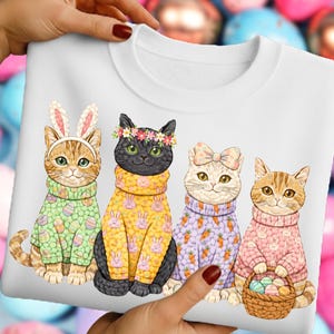 May include: White sweatshirt with a design of four cartoon cats in sweaters. One cat wears bunny ears, another a flower crown, and a third a bow. The cats' sweaters have Easter egg, carrot, and bunny patterns. The last cat holds a basket of Easter eggs.