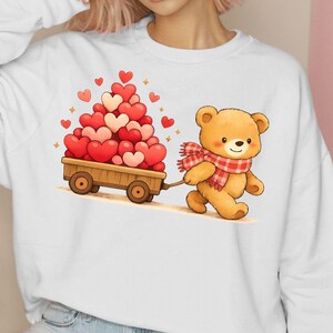May include: A white sweatshirt with a cartoon illustration of a teddy bear pulling a wooden cart filled with heart-shaped balloons. The bear wears a red and white checked scarf. The hearts are red and pink.
