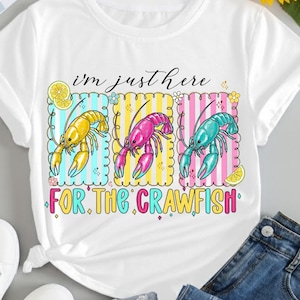 Könnte beinhalten: Weißes T-Shirt mit einer bunten Grafik von drei Krebsen in Gelb, Pink und Türkis. Der Text "I'm just here for the crawfish" steht unter den Krebsen. Das Design umfasst Zitronenscheiben und florale Akzente.