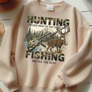 May include: A beige sweatshirt with a graphic featuring a rifle, a fish, and a deer. The words "HUNTING" and "FISHING" are printed in camouflage-style letters, with the phrases "SOLVED MOST OF THE PROBLEMS" and "SOLVES THE REST" below.