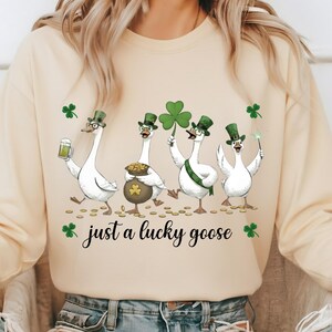 Puede incluir: Una sudadera color crema con un dise&ntilde;o del D&iacute;a de San Patricio. El gr&aacute;fico muestra cuatro gansos blancos con sombreros verdes, sosteniendo una cerveza, un pote de oro, un tr&eacute;bol y una varita m&aacute;gica. El texto dice "just a lucky goose."