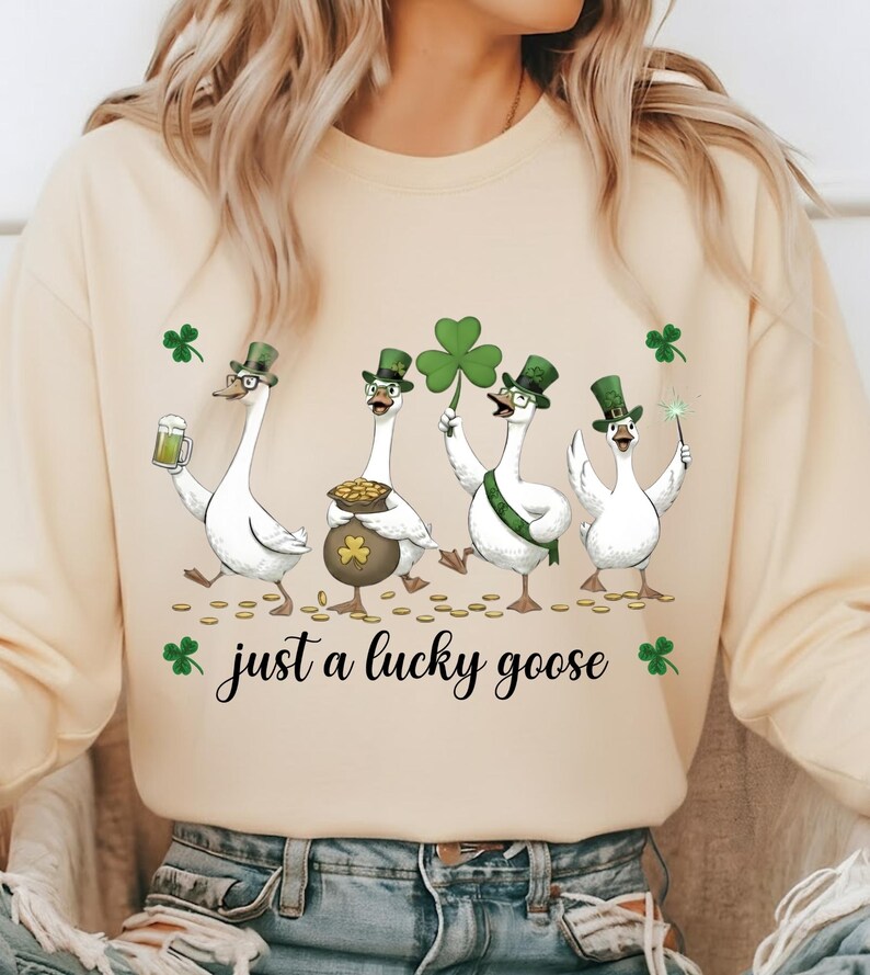 Puede incluir: Una sudadera color crema con un dise&ntilde;o del D&iacute;a de San Patricio. El gr&aacute;fico muestra cuatro gansos blancos con sombreros verdes, sosteniendo una cerveza, un pote de oro, un tr&eacute;bol y una varita m&aacute;gica. El texto dice "just a lucky goose."