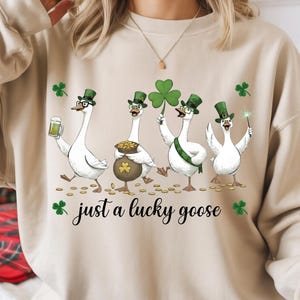 Puede incluir: Sudadera beige con cuatro gansos de dibujos animados celebrando el D&iacute;a de San Patricio. Llevan sombreros verdes y gafas. Uno sostiene una cerveza, otro una olla de oro, un tercero un tr&eacute;bol y el &uacute;ltimo una varita. El texto dice "just a lucky goose".