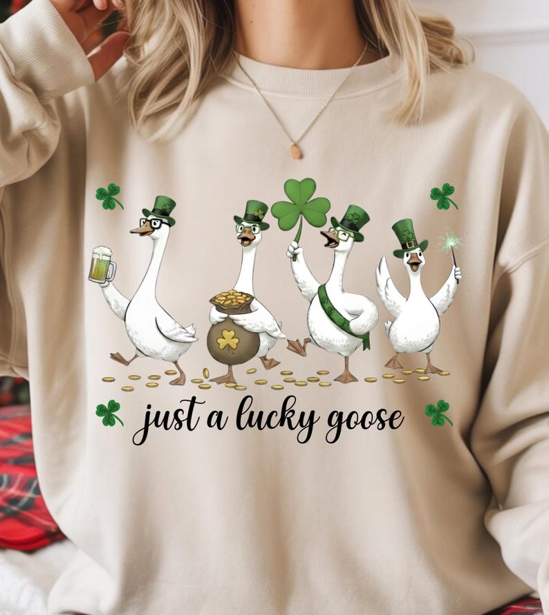 Puede incluir: Sudadera beige con cuatro gansos de dibujos animados celebrando el D&iacute;a de San Patricio. Llevan sombreros verdes y gafas. Uno sostiene una cerveza, otro una olla de oro, un tercero un tr&eacute;bol y el &uacute;ltimo una varita. El texto dice "just a lucky goose".