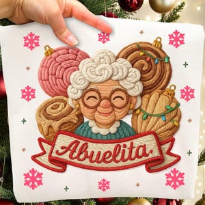 May include: A white square pillow with a festive design featuring a smiling cartoon grandmother with glasses, surrounded by decorative ornaments and snowflakes. The word "Abuelita" is written on a banner.