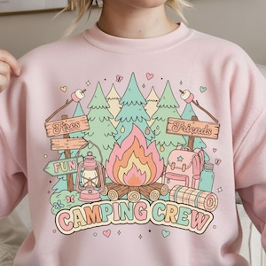 以下が含まれることがあります： ピンク色のスウェットシャツ。キャンプをテーマにしたカラフルなグラフィックが特徴です。デザインには、キャンプファイヤー、木、ランタン、バックパック、そして「Fires」、「Friends」、「Fun」、「Camping Crew」という言葉が含まれています。グラフィックはパステルカラーの漫画スタイルです。