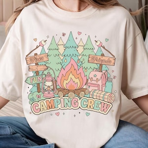 以下が含まれることがあります： クリーム色のTシャツ。キャンプをテーマにしたカラフルなグラフィックが特徴です。「Camping Crew」、「Fires」、「Fun」、「Friends」の文字と、キャンプファイヤー、木、ランタン、バックパックが描かれています。パステルカラーです。