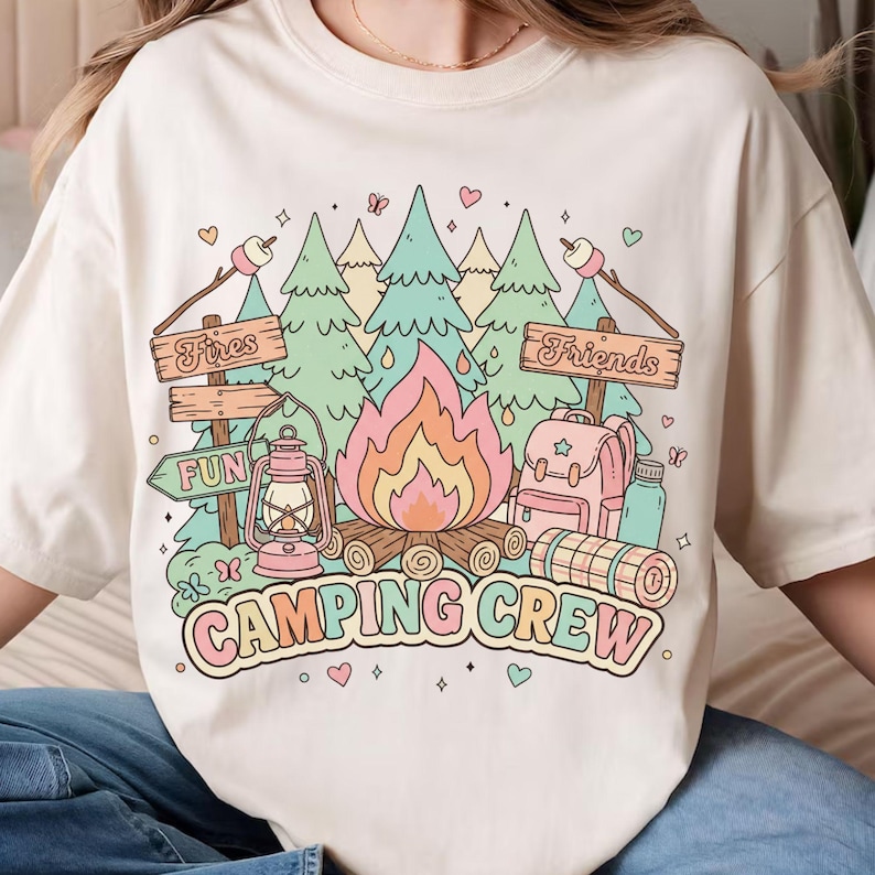 以下が含まれることがあります： クリーム色のTシャツ。キャンプをテーマにしたカラフルなグラフィックが特徴です。「Camping Crew」、「Fires」、「Fun」、「Friends」の文字と、キャンプファイヤー、木、ランタン、バックパックが描かれています。パステルカラーです。