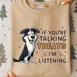 May include: A beige sweatshirt with a cartoon border collie illustration. The dog is black and white with a smiling face. Text on the front reads "IF YOU'RE TALKING TREATS I'M LISTENING". The sweatshirt has a round neck.