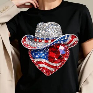 May include: Black t-shirt featuring a rhinestone-studded cowboy hat and a heart-shaped American flag design. The hat has a silver crown, red and blue brim, and the heart has a large red gem.