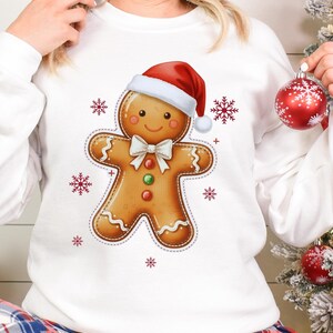 May include: White sweatshirt featuring a gingerbread man wearing a Santa hat and bow tie. The gingerbread man has a smiling face and is decorated with red, green, and orange details. Red snowflakes are scattered around the gingerbread man.