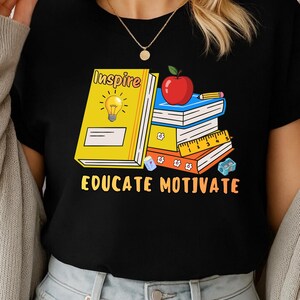 May include: Black t-shirt featuring a graphic of books, an apple, and a pencil. The text on the shirt reads "Inspire" and "Educate Motivate". The design includes a yellow book with a lightbulb.
