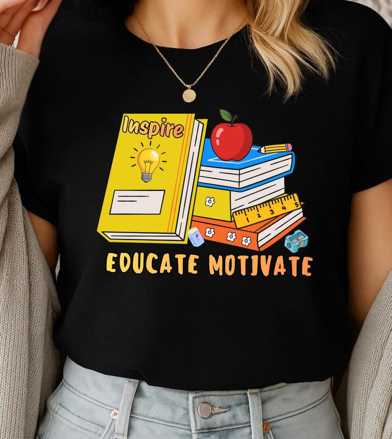 May include: Black t-shirt featuring a graphic of books, an apple, and a pencil. The text on the shirt reads "Inspire" and "Educate Motivate". The design includes a yellow book with a lightbulb.