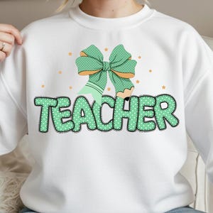 May include: White sweatshirt with a green bow and pencil graphic. The word "TEACHER" is printed in green, dotted letters. Small orange stars are scattered around the graphic.