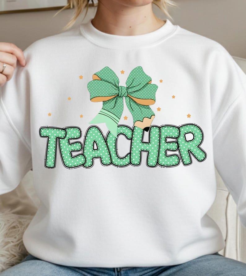 May include: White sweatshirt with a green bow and pencil graphic. The word "TEACHER" is printed in green, dotted letters. Small orange stars are scattered around the graphic.