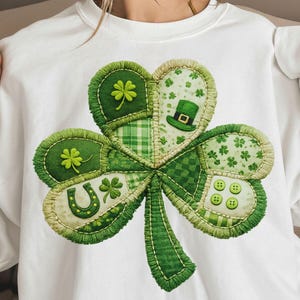 Może przedstawiać: Biała bluza z dużym, szczegółowym wzorem koniczyny. Koniczyna składa się z różnych zielonych tkanin, w tym wzorów w kratę i kropki, z podkową, guzikami i kapeluszem krasnoludka. Wzór jest zszyty kontrastującą nicią.