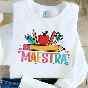 May include: White sweatshirt with a colorful graphic featuring school supplies like pencils, an apple, ruler, and scissors. The word "MAESTRA" is written in a playful, multi-colored font, perfect for teachers.