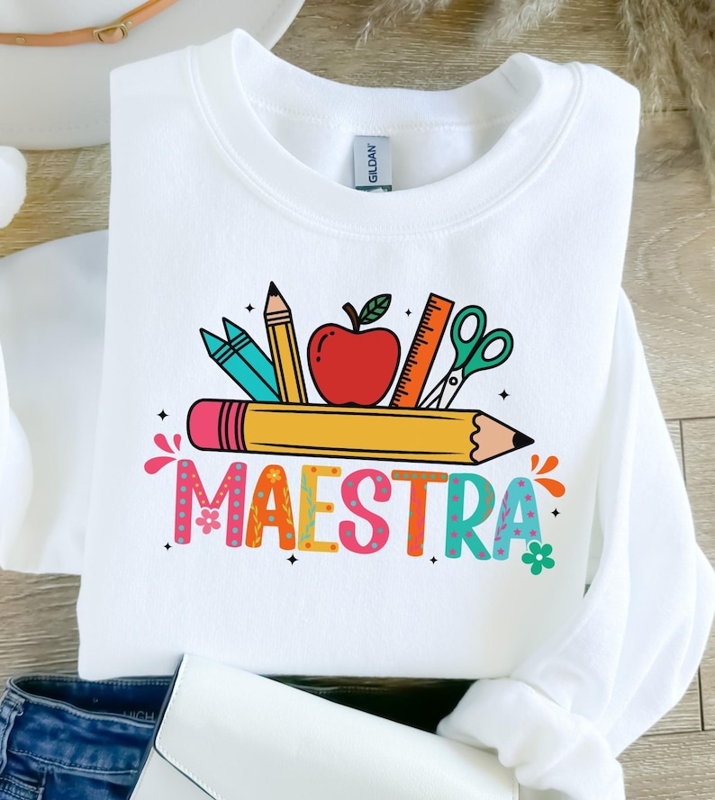 May include: White sweatshirt with a colorful graphic featuring school supplies like pencils, an apple, ruler, and scissors. The word "MAESTRA" is written in a playful, multi-colored font, perfect for teachers.