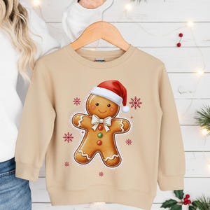 May include: A tan-colored sweatshirt featuring a gingerbread man graphic wearing a Santa hat and bow tie. Red and white snowflakes surround the gingerbread man. The sweatshirt is hanging on a wooden hanger.