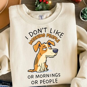 May include: Cream-coloured sweatshirt featuring the text "I DON'T LIKE MORNING PEOPLE OR MORNINGS OR PEOPLE" above a cartoon dog with a grumpy expression. The dog is brown and white with a black nose and is sitting down.