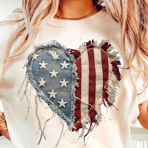 Op de afbeelding: Crèmekleurig T-shirt met een hartontwerp dat in twee helften is verdeeld. De ene helft is spijkerblauw met witte sterren, de andere heeft rood-witte strepen. Het hart heeft een gerafelde, versleten rand.