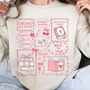 May include: Beige sweatshirt with a pink and white graphic design featuring books, hearts, and the phrases "ONE BED ALL THE INN", "enemies to lovers", "grumpy x sunshine", "just one more chapter", and "born to read".