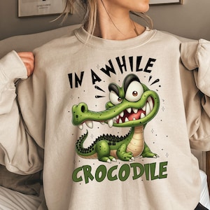 K&ouml;nnte beinhalten: Beiges Sweatshirt mit einem Cartoon-Krokodil-Design und dem Text "IN A WHILE CROCODILE". Das Krokodil ist gr&uuml;n mit wei&szlig;en Z&auml;hnen und einem schwarzen R&uuml;cken. Das Sweatshirt hat lange &Auml;rmel und einen Rundhalsausschnitt.