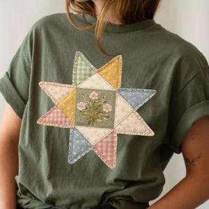 May include: An olive green t-shirt featuring a patchwork star design. The star is composed of various fabric patterns and colors, including floral embroidery, gingham, and pastel hues. The shirt has short sleeves and a relaxed fit.