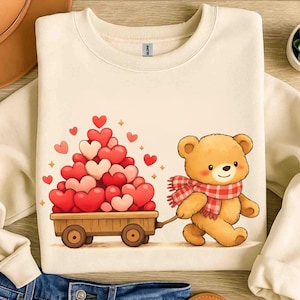 May include: Cream-coloured sweatshirt with a cartoon teddy bear pulling a wooden cart filled with heart-shaped balloons. The bear wears a red and white checked scarf. The cart is filled with red and pink hearts.