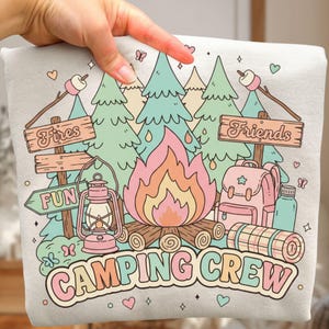 以下が含まれることがあります： キャンプをテーマにしたカラフルなグラフィックがプリントされたライトグレーのTシャツ。デザインには、キャンプファイヤー、木、バックパック、「Camping Crew」の文字が含まれています。「Fires」、「Fun」、「Friends」の文字も含まれています。