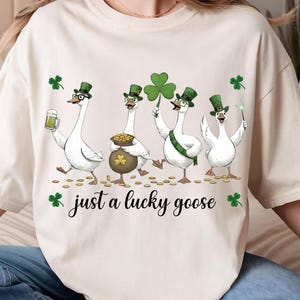 Puede incluir: Camiseta color crema con una ilustraci&oacute;n caprichosa de cuatro gansos blancos vestidos para el D&iacute;a de San Patricio. Cada ganso lleva un sombrero verde y sostiene objetos festivos. El texto "just a lucky goose" est&aacute; impreso debajo.