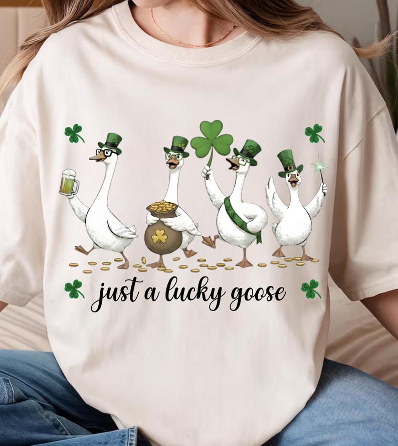 Puede incluir: Camiseta color crema con una ilustraci&oacute;n caprichosa de cuatro gansos blancos vestidos para el D&iacute;a de San Patricio. Cada ganso lleva un sombrero verde y sostiene objetos festivos. El texto "just a lucky goose" est&aacute; impreso debajo.