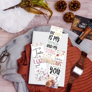 May include: A collection of floral bookmarks with inspirational quotes, resting on a white book. The bookmarks feature phrases like "The Lord is My Strength" and "Taste and see the Lord." The scene includes a watch, makeup, and a sweater.