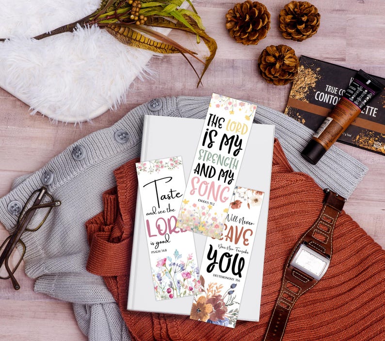 May include: A collection of floral bookmarks with inspirational quotes, resting on a white book. The bookmarks feature phrases like "The Lord is My Strength" and "Taste and see the Lord." The scene includes a watch, makeup, and a sweater.