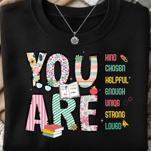 May include: Black sweatshirt with the words "YOU ARE" in colorful, floral, and striped designs. The words "Kind, Chosen, Helpful, Enough, Unique, Strong, Loved" are printed vertically. A silver necklace with a round pendant is visible.