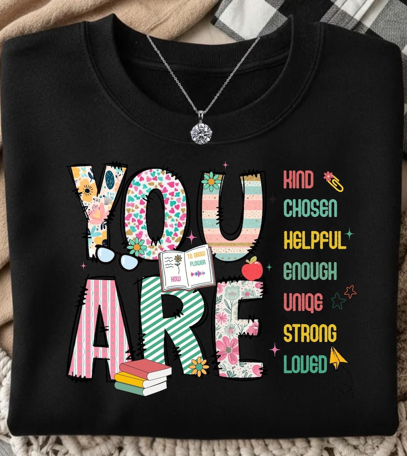 May include: Black sweatshirt with the words "YOU ARE" in colorful, floral, and striped designs. The words "Kind, Chosen, Helpful, Enough, Unique, Strong, Loved" are printed vertically. A silver necklace with a round pendant is visible.