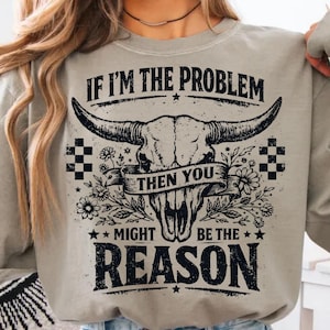 Pode incluir: Camisola cinza claro com um gráfico de caveira de touro, flores e o texto "IF I'M THE PROBLEM THEN YOU MIGHT BE THE REASON" em preto. O design inclui padrões quadriculados e estrelas.
