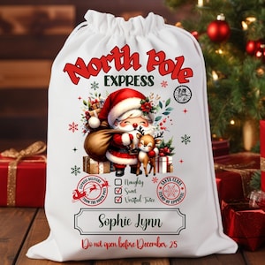 May include: A white drawstring Christmas gift bag with a festive illustration of Santa Claus and a reindeer. The bag features the text "North Pole Express" and "Sophie Lynn". The bag is decorated with snowflakes and gift boxes.