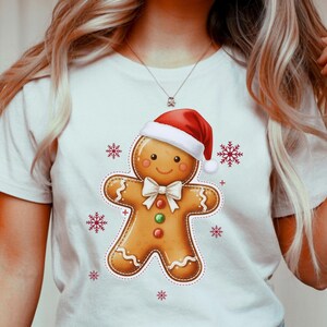 May include: White t-shirt featuring a gingerbread man wearing a Santa hat and a white bow tie. The gingerbread man has red, green, and orange buttons. Red snowflakes are scattered around the gingerbread man. The shirt is a festive holiday design.