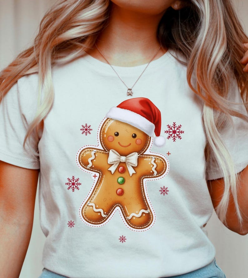 May include: White t-shirt featuring a gingerbread man wearing a Santa hat and a white bow tie. The gingerbread man has red, green, and orange buttons. Red snowflakes are scattered around the gingerbread man. The shirt is a festive holiday design.