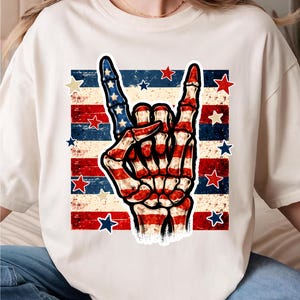 May include: Cream-colored t-shirt featuring a graphic of a skeleton hand making a rock and roll gesture, with red, white, and blue stripes and stars, reminiscent of the American flag. The design is centered on the shirt.