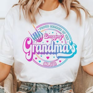 May include: White t-shirt with a colorful graphic that reads "Proud Member of the Bragging Grandma's Club." The design includes a skeleton hand making a rock-and-roll gesture, hearts, and a retro font.