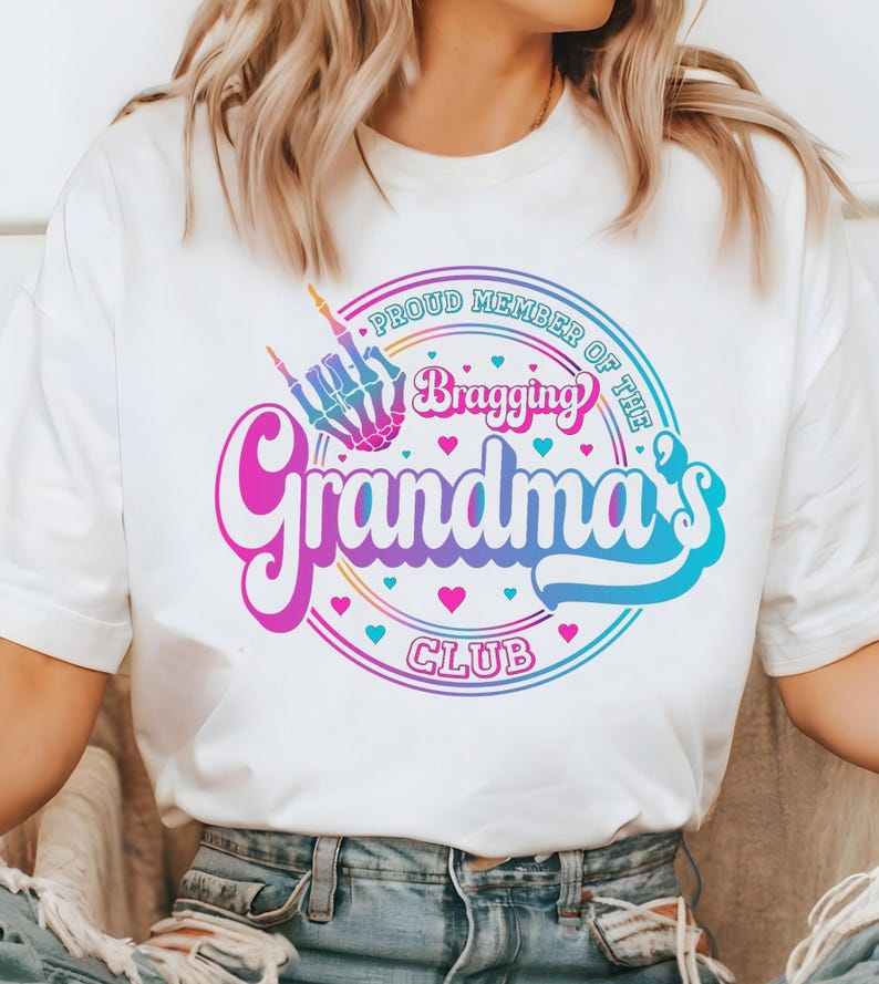 May include: White t-shirt with a colorful graphic that reads "Proud Member of the Bragging Grandma's Club." The design includes a skeleton hand making a rock-and-roll gesture, hearts, and a retro font.