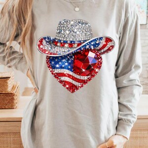 May include: A long-sleeved gray shirt featuring a patriotic design. The design includes a bedazzled cowboy hat and a heart-shaped American flag with a large red gem in the center. The hat and heart are covered in sparkling rhinestones.