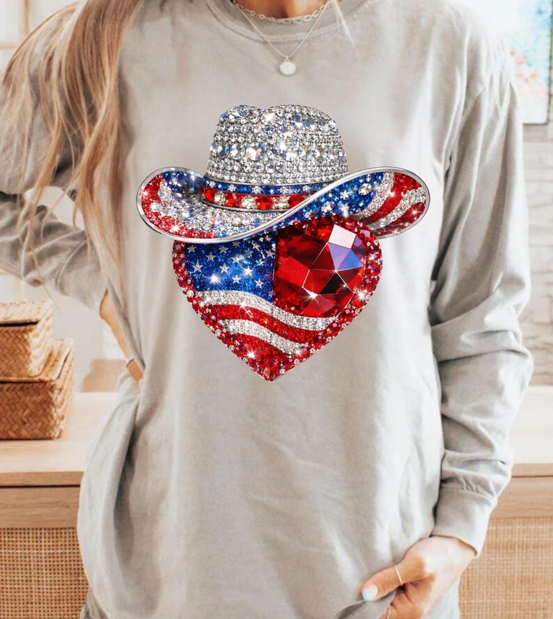 May include: A long-sleeved gray shirt featuring a patriotic design. The design includes a bedazzled cowboy hat and a heart-shaped American flag with a large red gem in the center. The hat and heart are covered in sparkling rhinestones.
