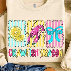 May include: A cream-coloured sweatshirt with a vibrant graphic. It displays a lemon slice, a pink crawfish, and a blue bow, each set against striped backgrounds. The text "CRAWFISH SEASON" is written below the illustrations.