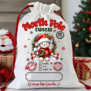 May include: White drawstring Christmas gift bag with red accents. The bag features a teddy bear wearing a Santa hat and sweater, surrounded by a wreath. Text includes "North Pole Express" and "Do not open before December 25."