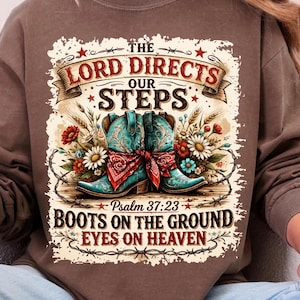 May include: Brown sweatshirt with a graphic design featuring turquoise cowboy boots, a red bandana, and floral accents. The text reads "The Lord Directs Our Steps" and "Boots on the Ground Eyes on Heaven."